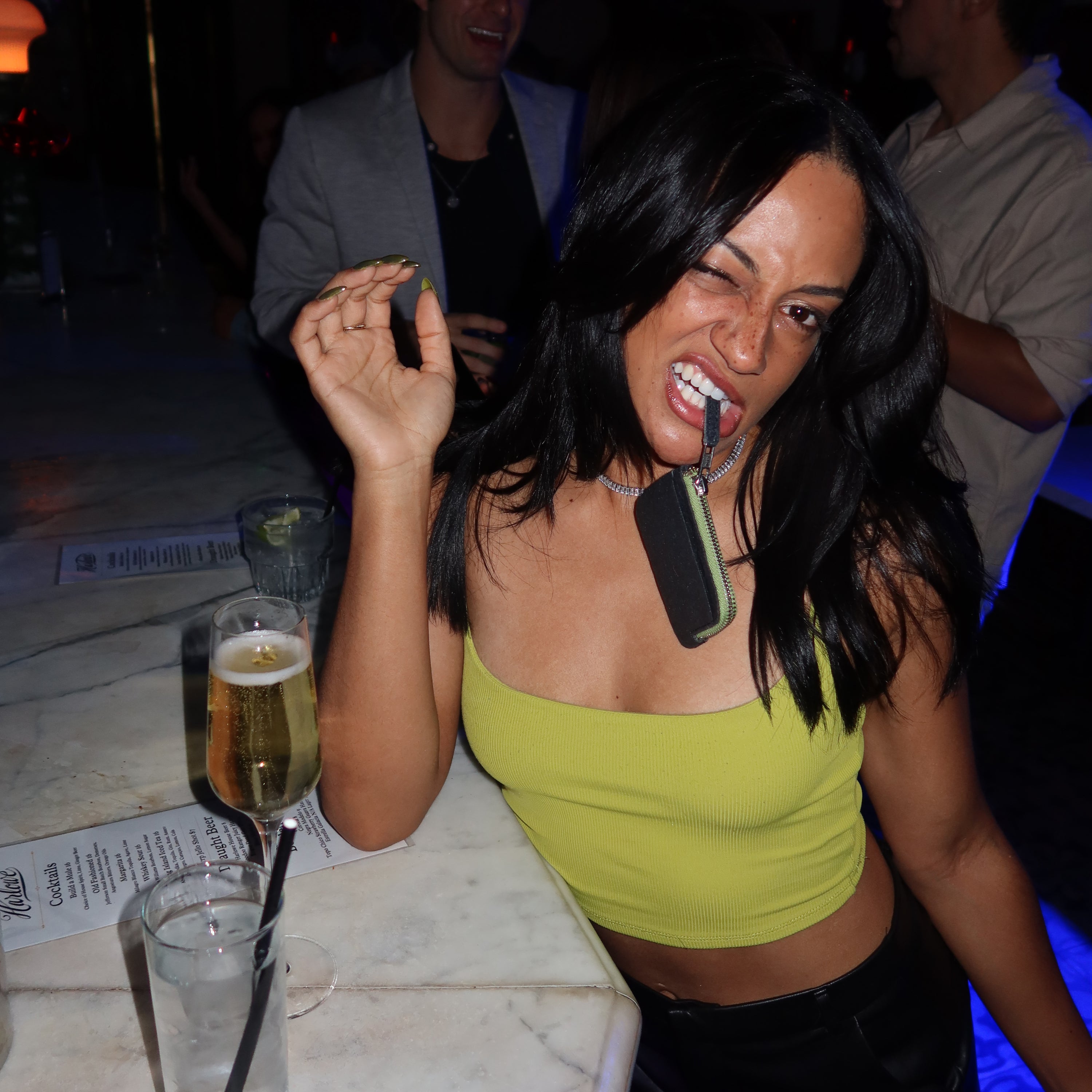 A woman poses playfully at a bar, winking and holding a Ground “The Zippered” wallet in her mouth by the zipper. She is wearing a bright yellow cropped tank top, with her black hair cascading down her shoulders. On the marble bar counter, there is a glass of champagne and a cocktail with a straw. The background is dimly lit with people socializing, and the scene suggests a fun night out.