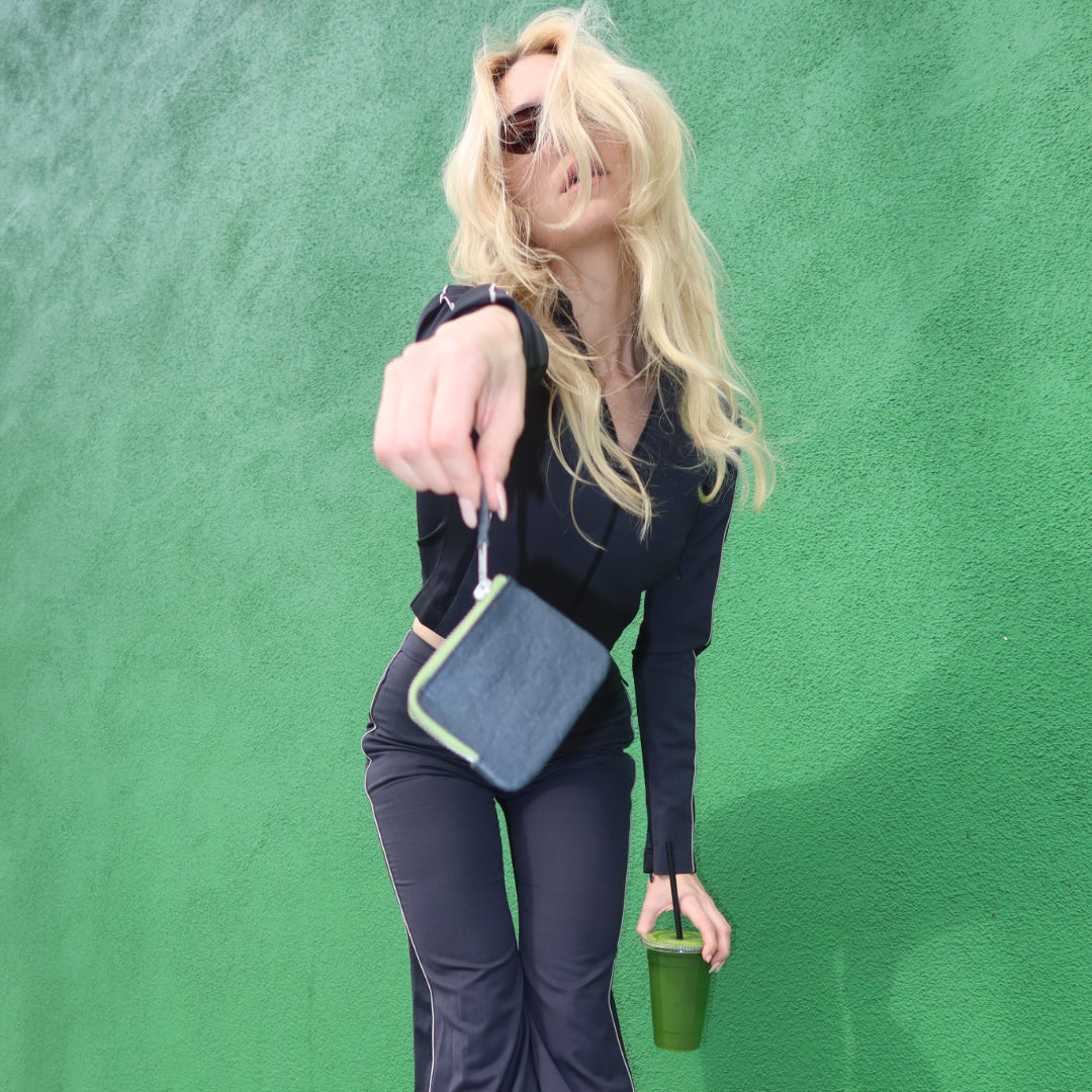 A woman with long blonde hair, wearing sunglasses, playfully leans forward while holding a Ground “The Zippered” wallet in one hand and a green smoothie in the other. She is dressed in a sleek dark outfit with piping details along the sides, set against a vibrant green wall. Her hair is windswept, adding movement and energy to the image. The bold contrast between her dark outfit and the bright green background, along with the playful attitude in her pose, creates a dynamic and stylish scene.